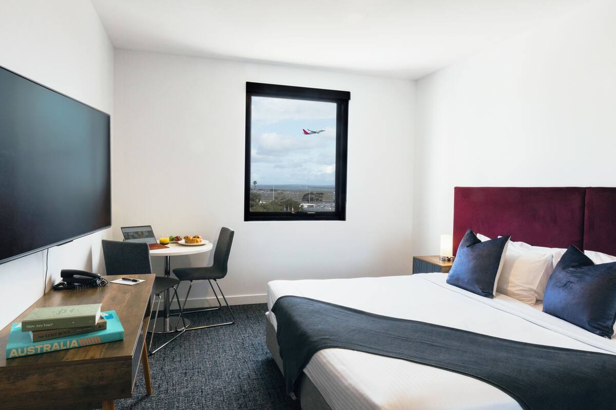 Ett hotellrum med en säng, ett skrivbord med en bärbar dator, en stol, ett fönster med utsikt över ett flygplan och en platt-TV.