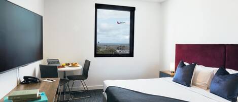 A hotel room with a bed, a desk with a laptop, a chair, a window with a view of an airplane, and a flat-screen TV.