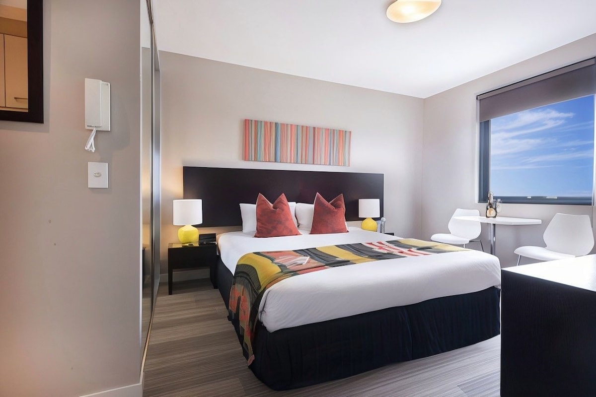 A hotel room with a large bed, bedside lamps, a small table, and a view of the sea.