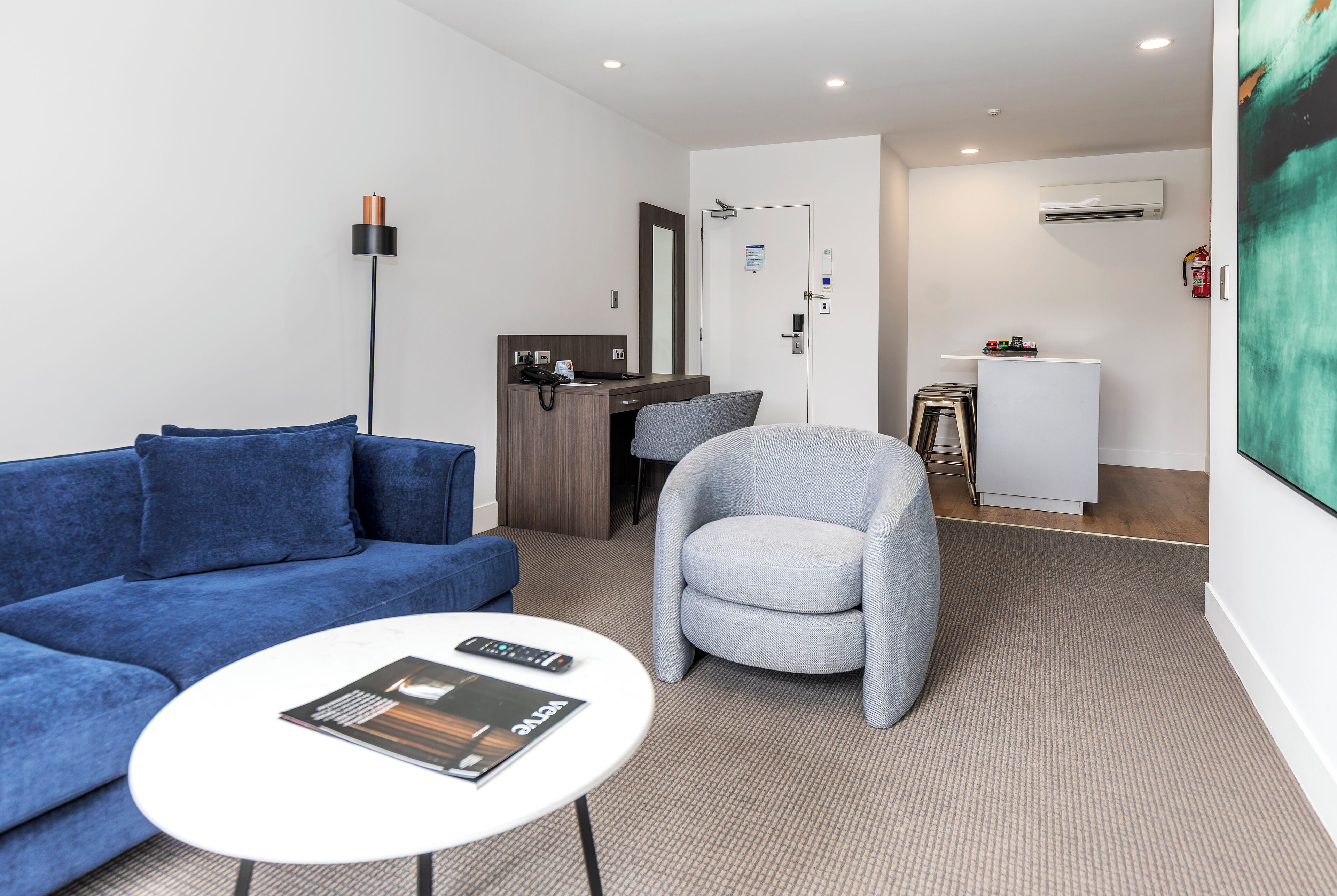 Ein modernes Hotelzimmer mit einem blauen Sofa, einem grauen Sessel, einem kleinen weißen Tisch und einem Schreibtisch mit einem Computer.