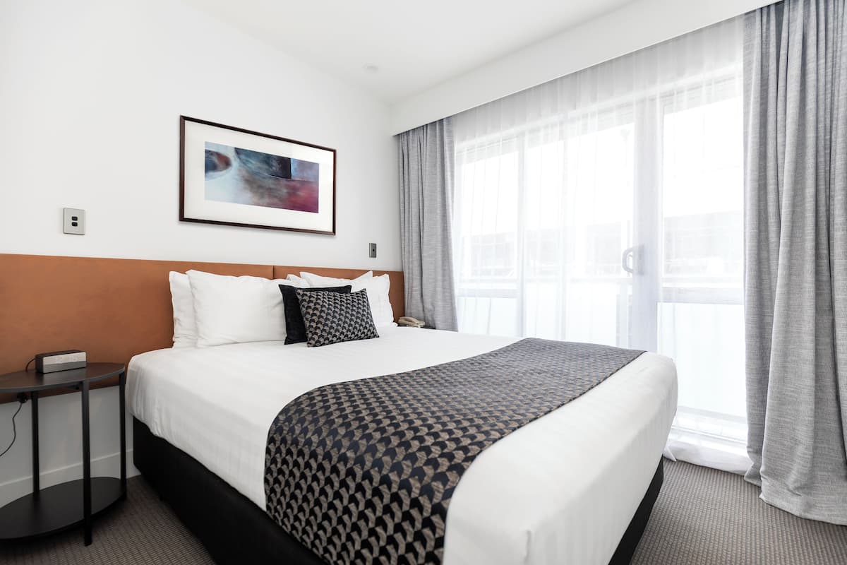 A hotel room with a large bed, a bedside table, a framed picture on the wall, and a window with curtains.