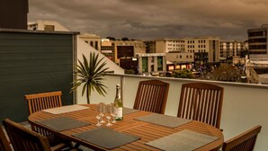 A wooden table set for four with a bottle of champagne, two glasses, and a plant on a balcony overlooking a cityscape. - Quest Newmarket (Auckland)