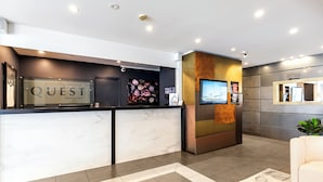 A modern hotel reception area with a marble counter, a large floral artwork, a flat-screen TV, and a seating area with a patterned rug. - Quest Newmarket (Auckland)