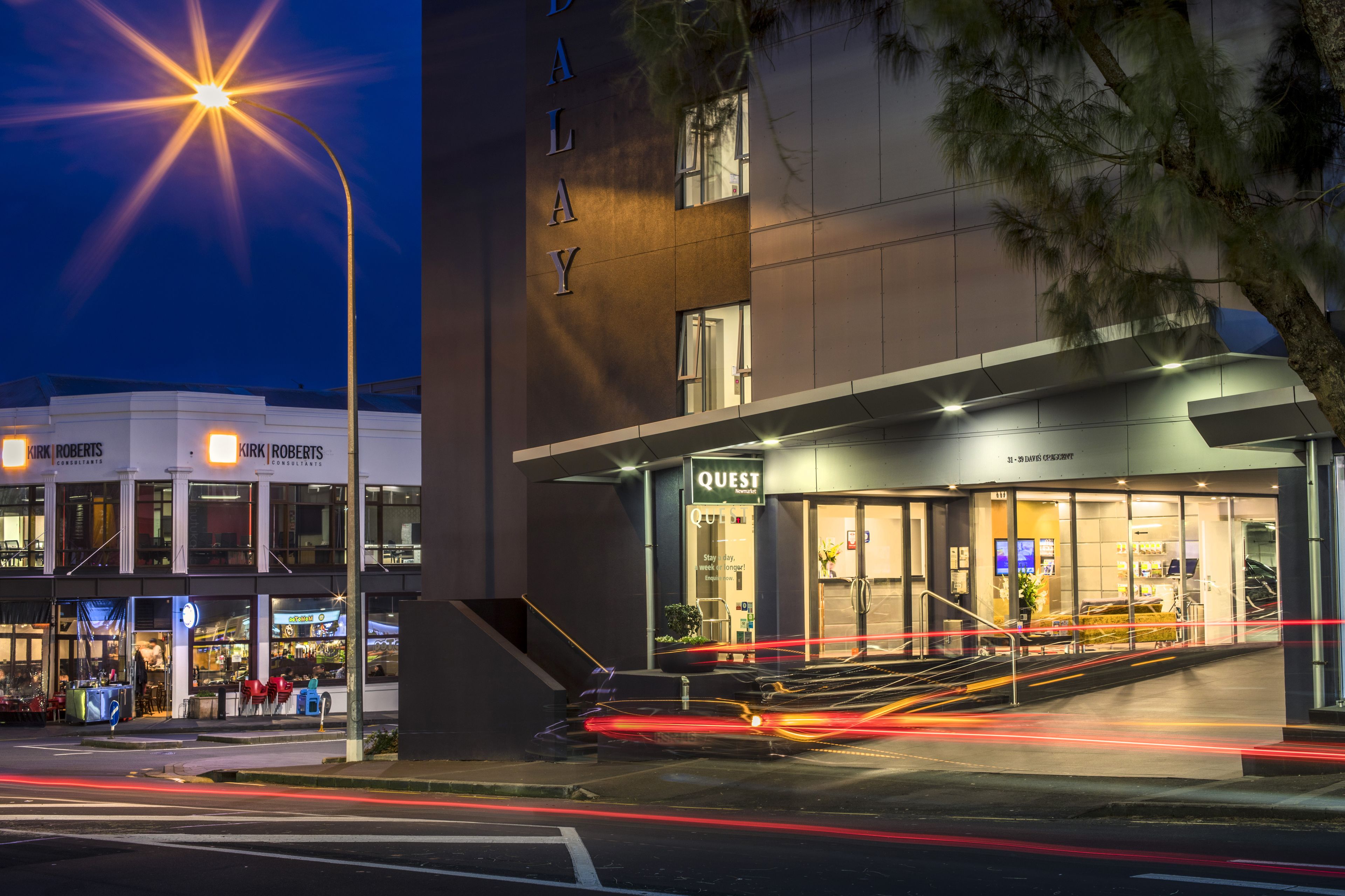 A modern building with a 'Quest' sign, a staircase leading to the entrance, and a street with light trails.