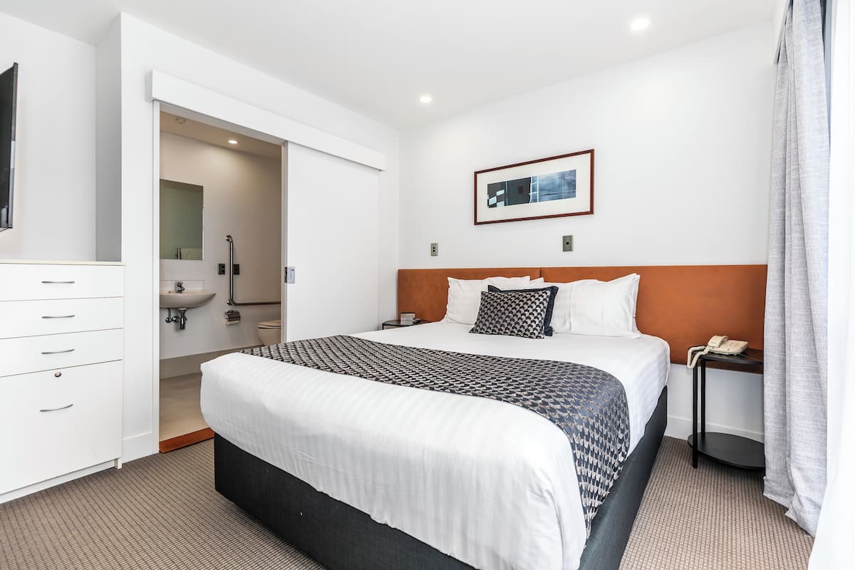 A hotel room with a large bed, a bedside table with a phone, a mirror, a bathroom with a sink and toilet, and a framed picture on the wall.