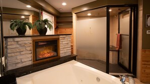 A modern bathroom with a large bathtub, a glass-enclosed shower, and a fireplace with two potted plants.