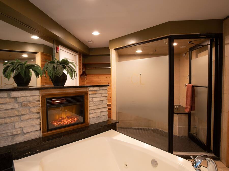 A modern bathroom with a large bathtub, a glass-enclosed shower, and a fireplace with two potted plants.