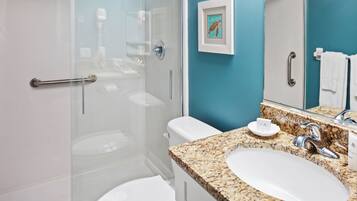 Une salle de bain dotée d’une douche à parois de verre, de toilettes blanches, d’un lavabo sur un plan de travail en granit et d’une œuvre d’art encadrée au mur.