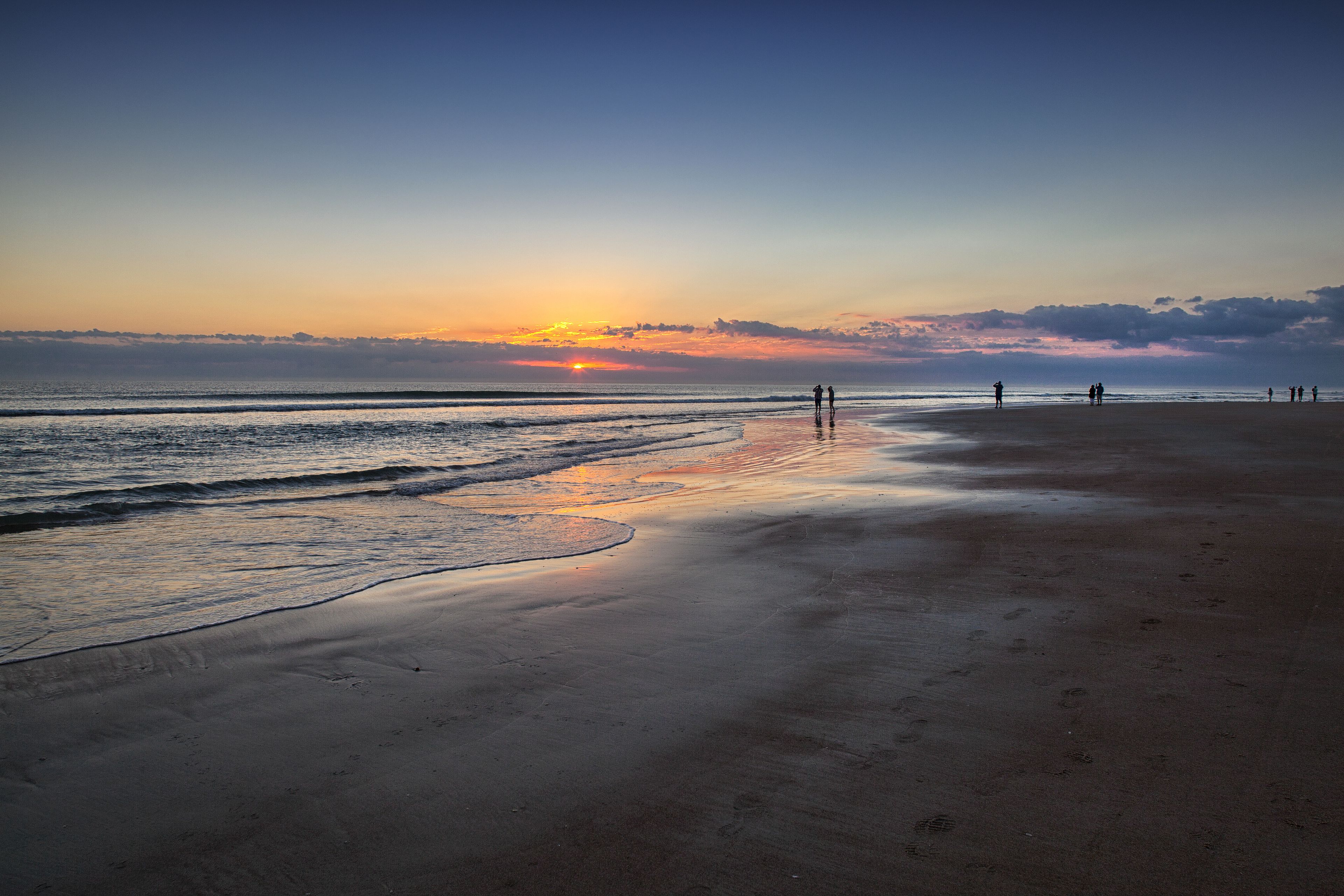 A beach with a sunset, calm waves, and a few people walking.