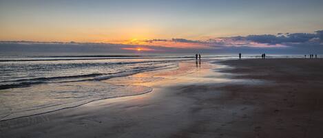 A beach with a sunset, calm waves, and a few people walking.