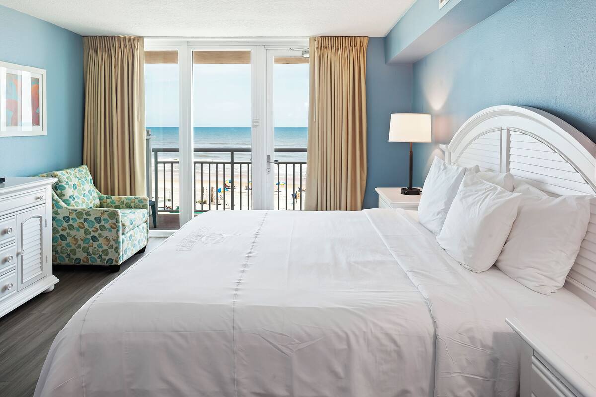 A hotel room with a large bed, a chair, a nightstand, and a view of the beach.