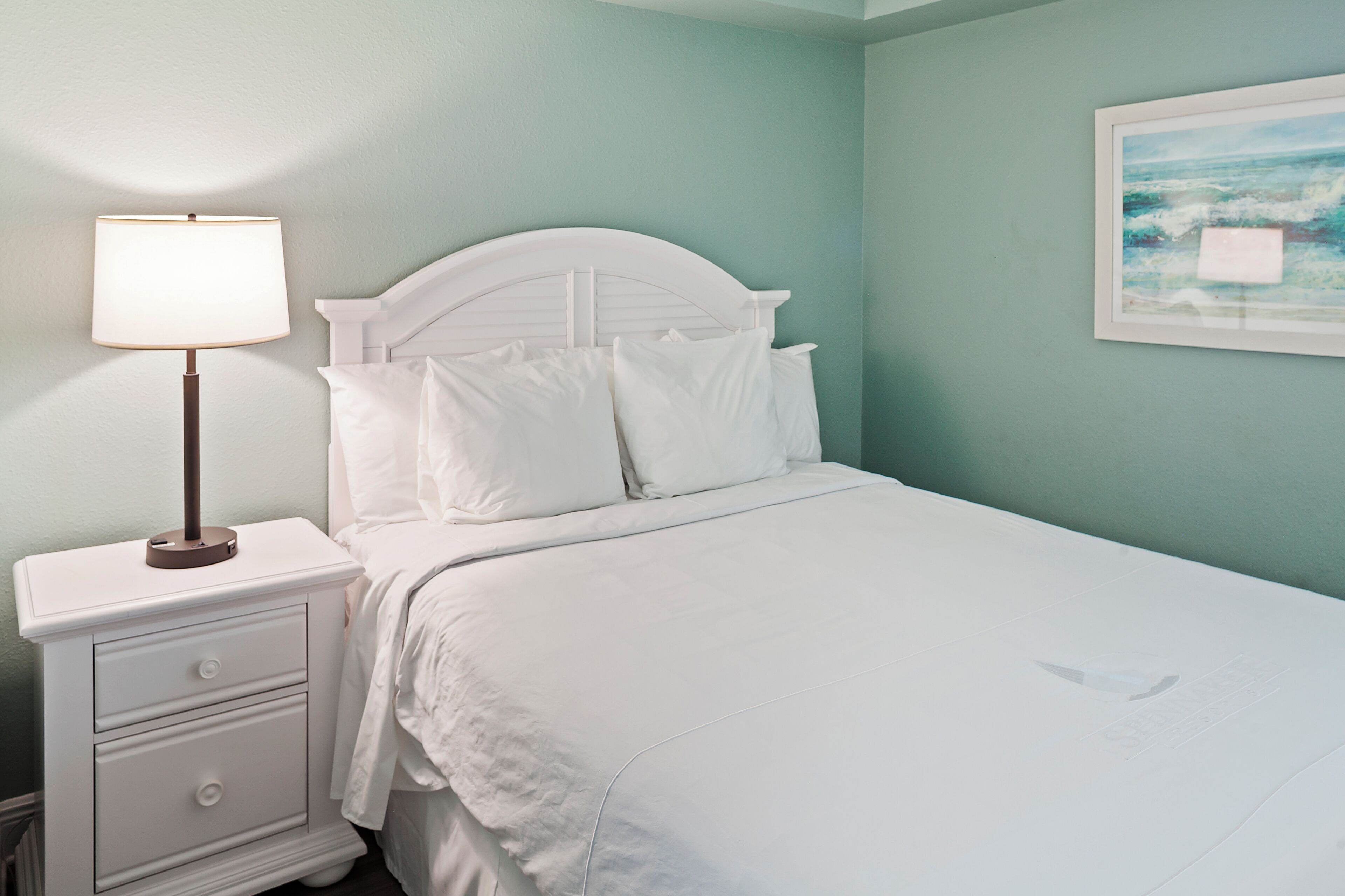 A neatly made bed with white linens, a bedside table with a lamp, and a framed picture on the wall.