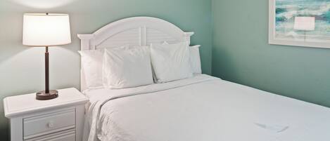 Una cama bien hecha con sábanas blancas, una mesita de noche con una lámpara y un cuadro en la pared.