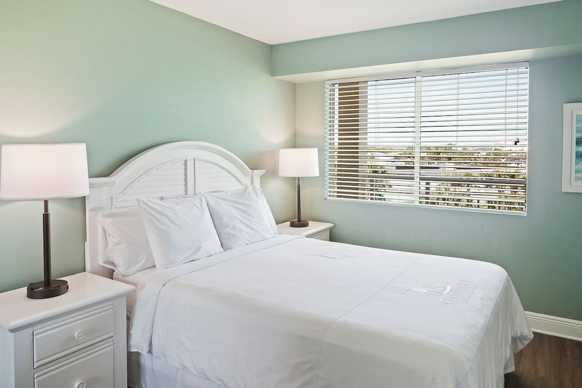 A neatly made bed with white bedding, two bedside tables with lamps, and a window with blinds.