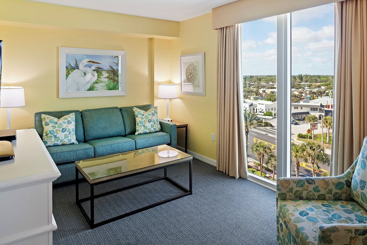 A hotel room with a green sofa, a coffee table, and a view of the city through large windows.