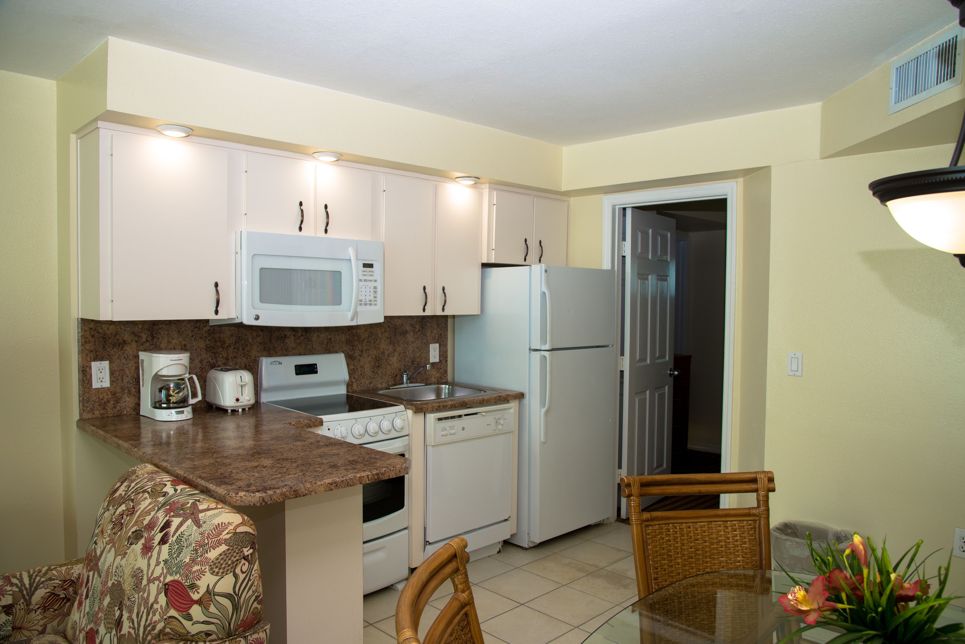 A compact kitchen with white appliances, a dining area with a glass table, and a wicker chair.