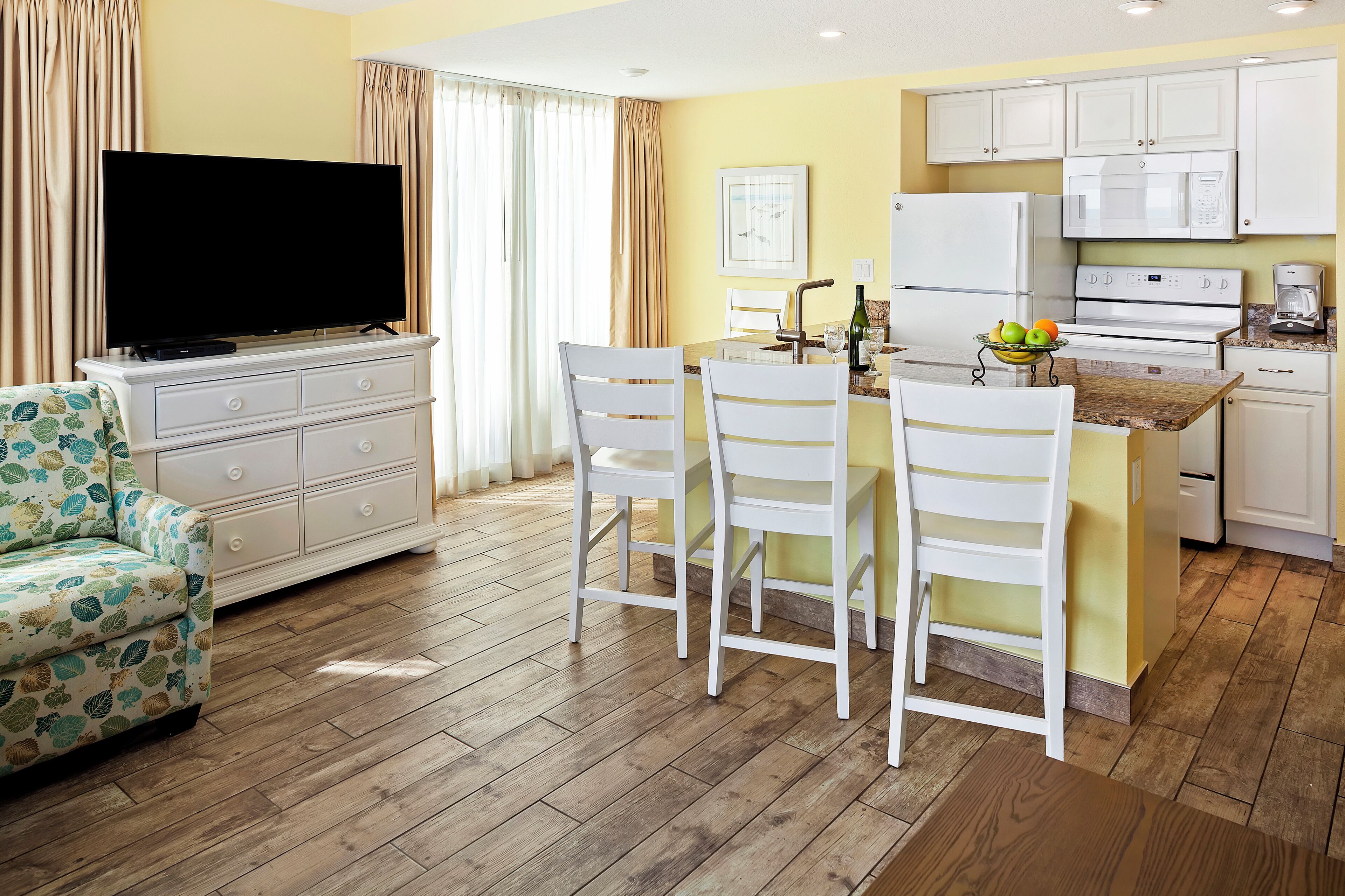 A modern kitchen with white cabinets, a dining area with a table and chairs, a flat-screen TV on a dresser, and a patterned armchair.