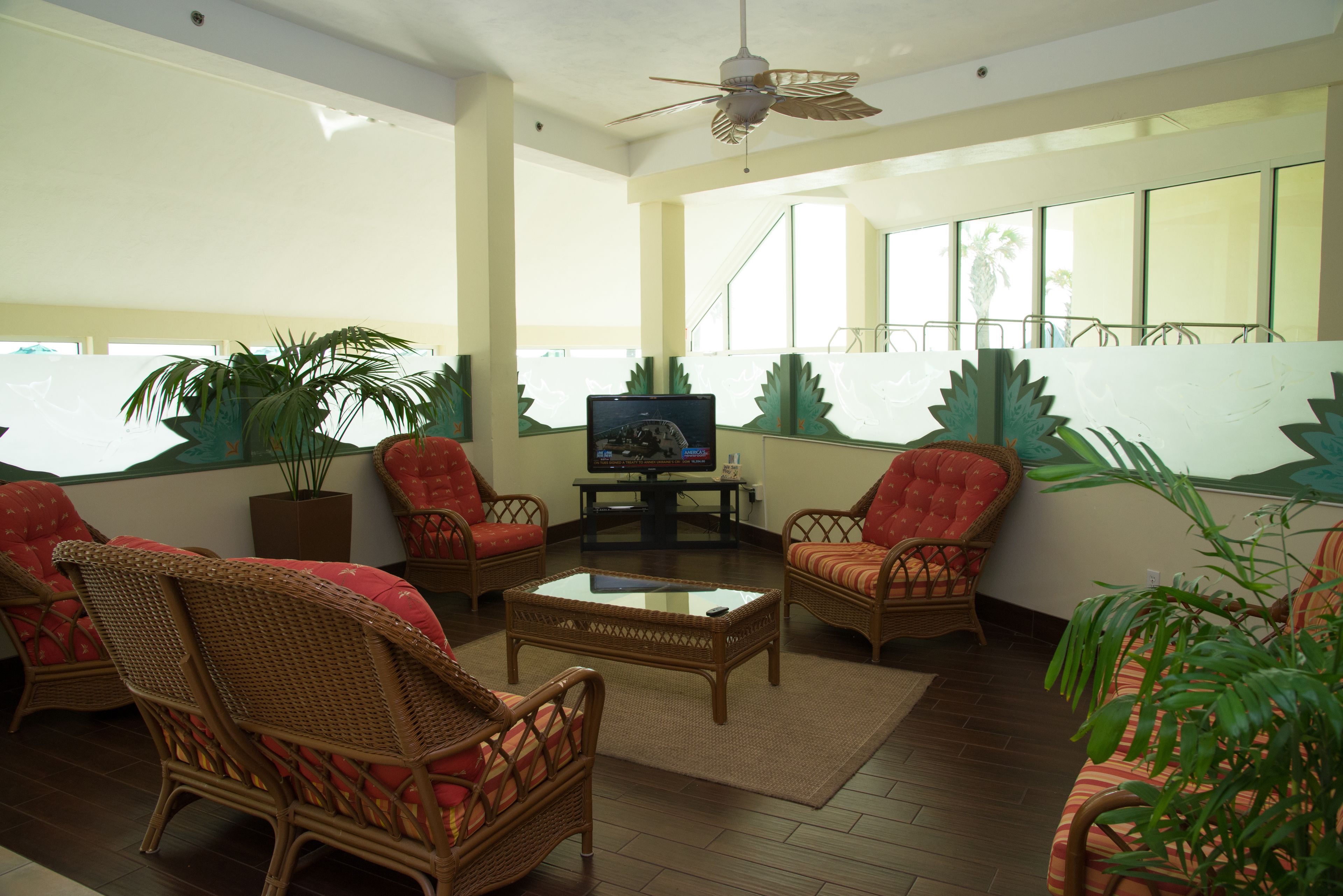 A spacious living area with wicker furniture, a television, and large windows offering a view of palm trees.