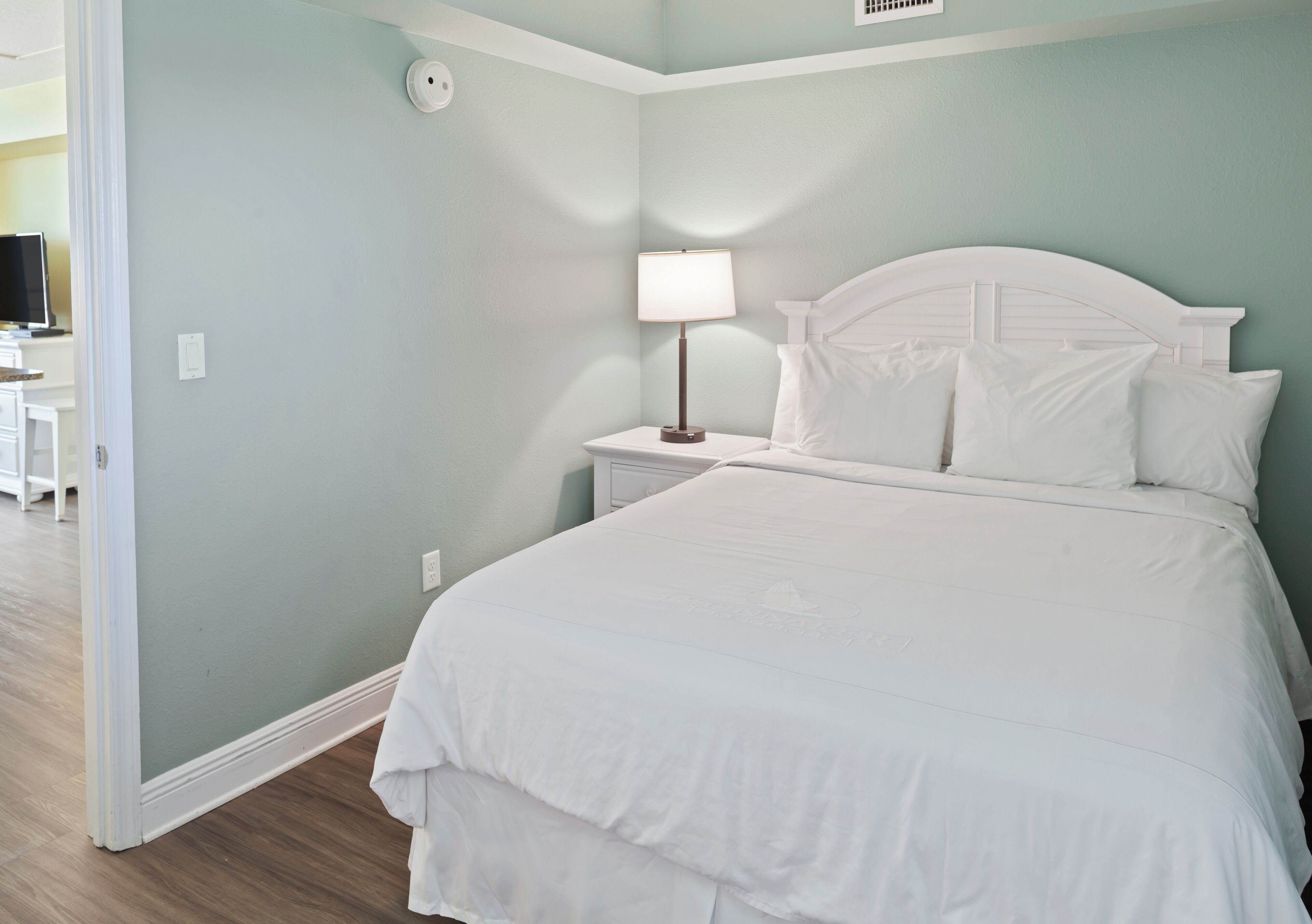 A bedroom with a white bed, a nightstand with a lamp, and a television in the background.