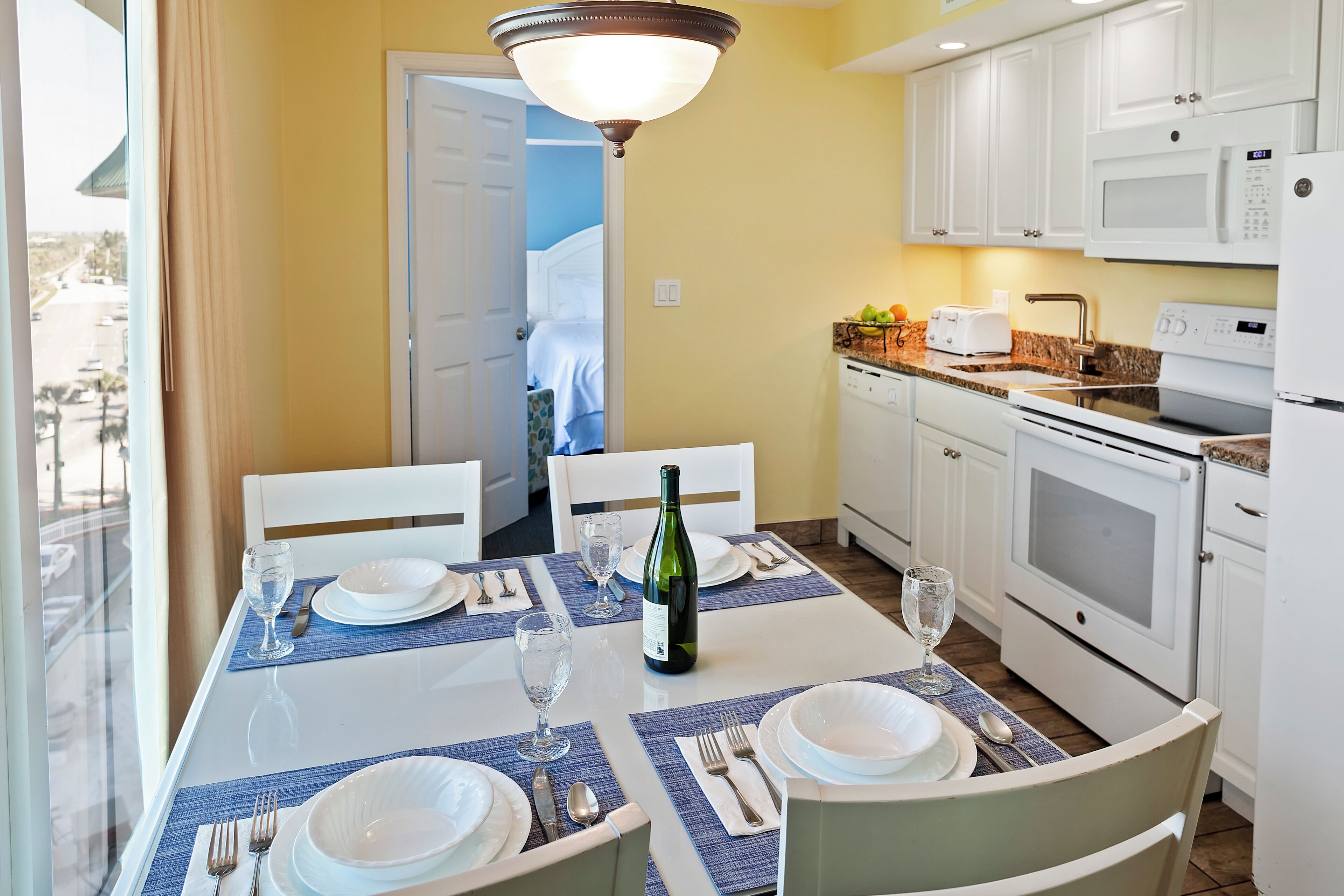 A dining table set for four with white plates, silverware, and a bottle of wine. A kitchen with white cabinets and appliances is visible in the background.