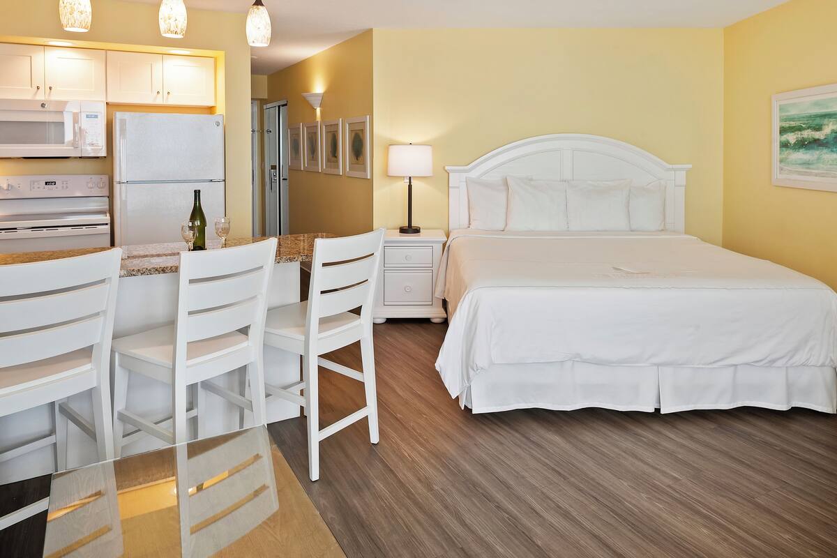 A hotel room with a bed, a desk with chairs, a refrigerator, a microwave, and a nightstand with a lamp.