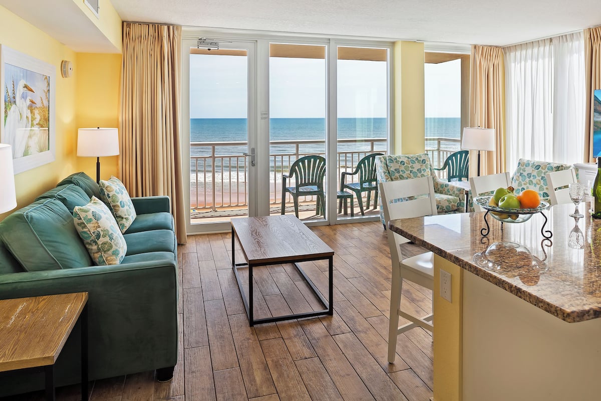 A room with a green sofa, a wooden coffee table, and a dining area with a fruit bowl and glassware. There are chairs and a view of the ocean through large glass doors.