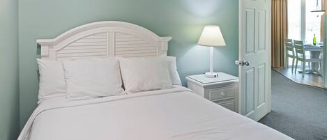 A neatly made bed with white bedding and a logo, a white nightstand with a lamp, and a door leading to a dining area.