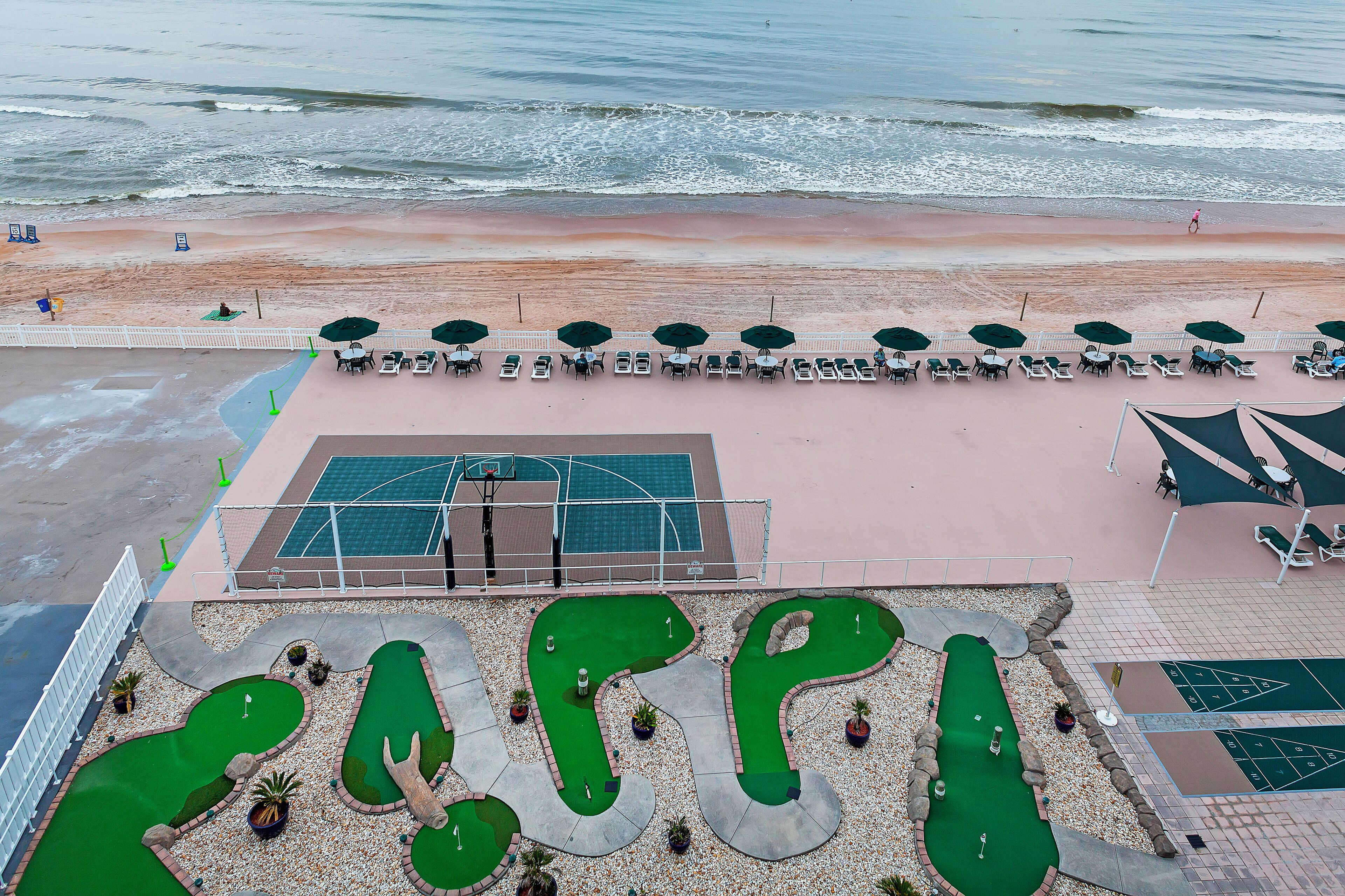 A beachside hotel with a pool table area, a mini-golf course, and a beach with umbrellas and lounge chairs.