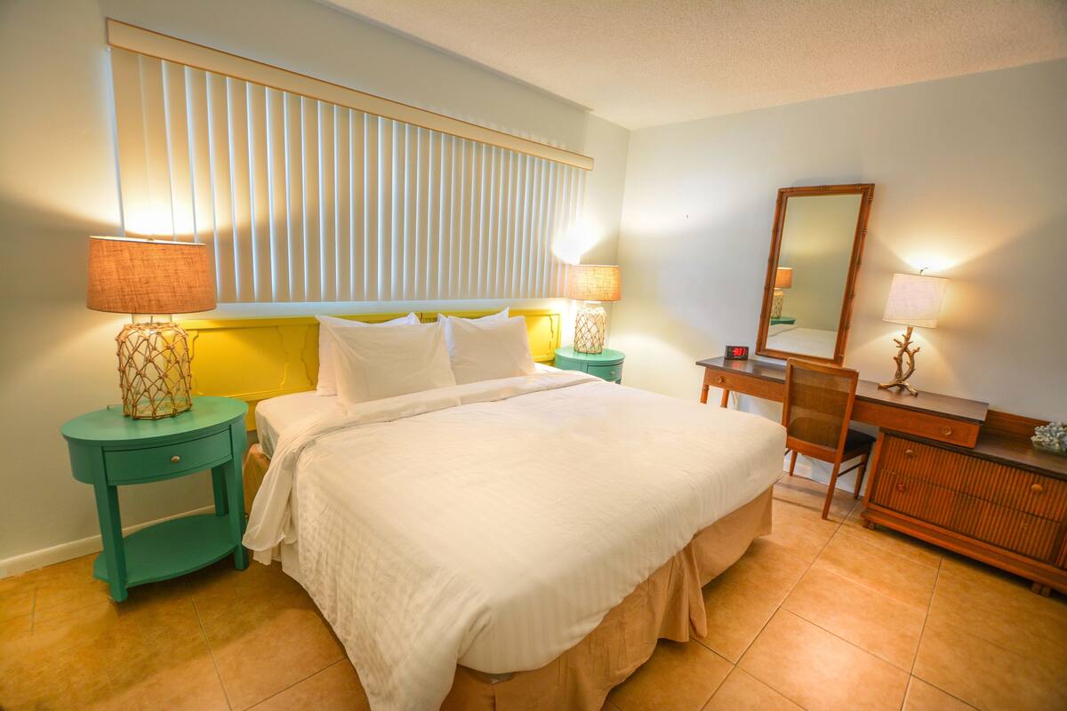Ein Hotelzimmer mit einem großen Bett, zwei Nachttischen mit Lampen, einer Kommode, einem Spiegel und einem Fenster mit Jalousien.