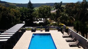Una piscina rectangular con sillas de descanso y un área de estar cubierta, rodeada de árboles y colinas al fondo.