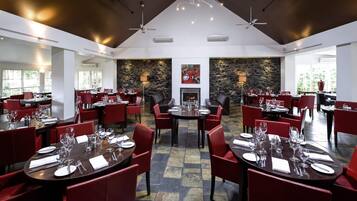 A spacious restaurant with multiple dining areas, red upholstered chairs, and a stone wall with a fireplace.