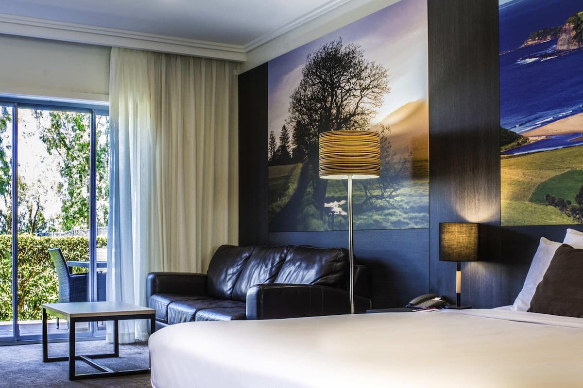A hotel room with a bed, a leather sofa, a table, a lamp, and a large window with curtains.
