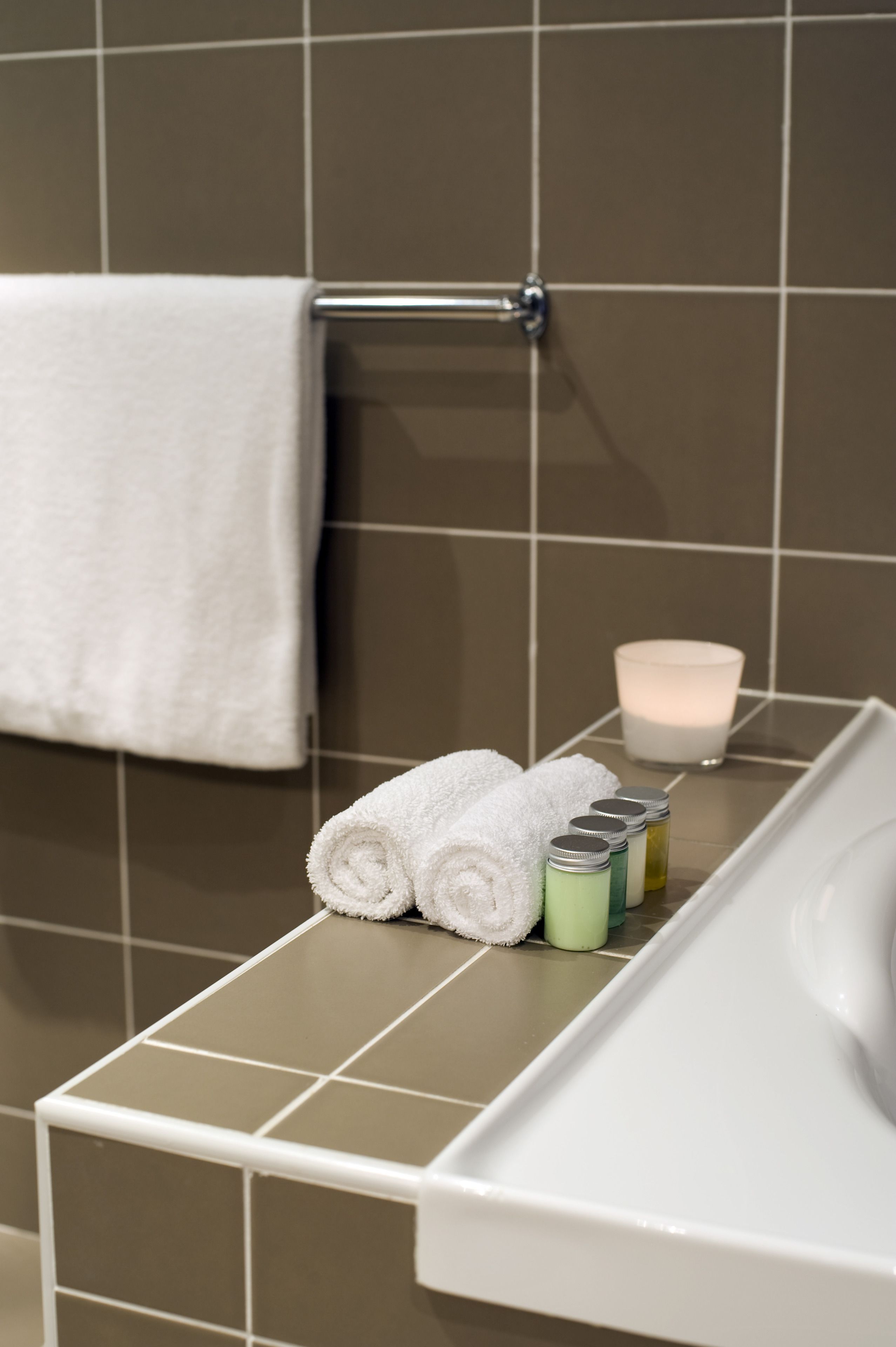 A bathroom with a white towel hanging on a rack, a rolled-up towel, and a set of toiletries on a tray.