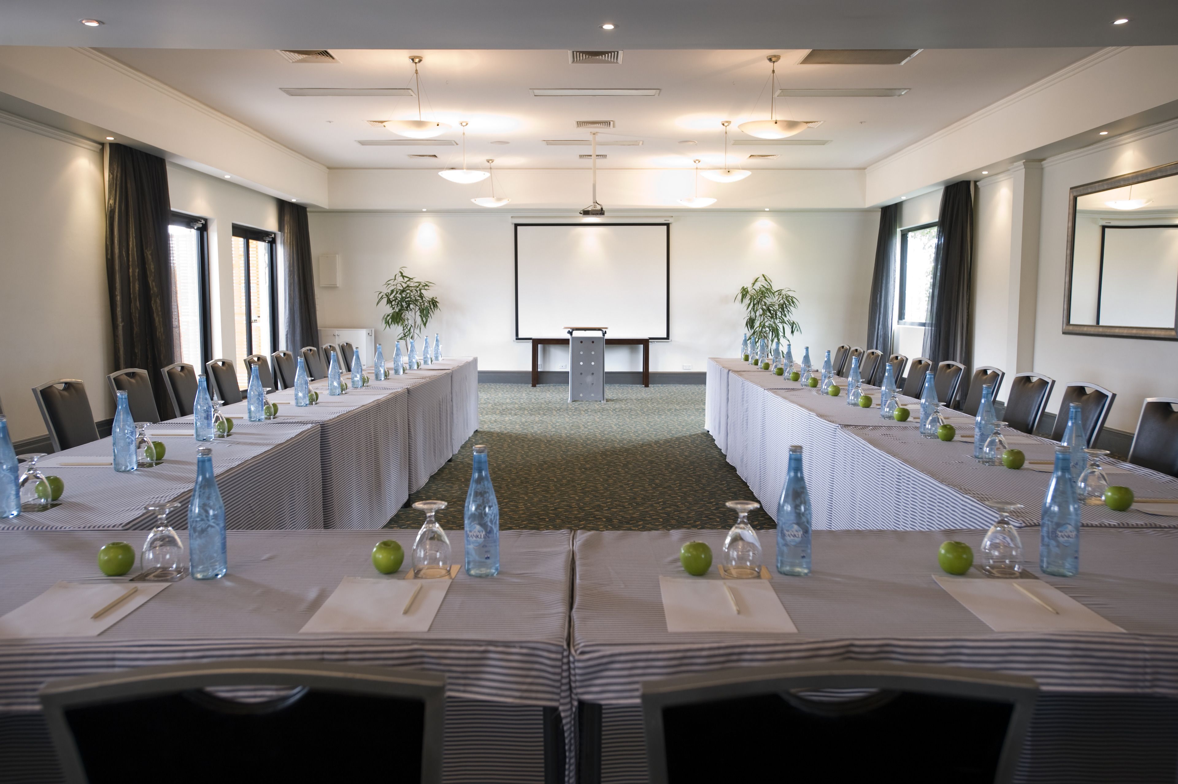 A conference room with a long table, chairs, a projector screen, and a large mirror on the wall.