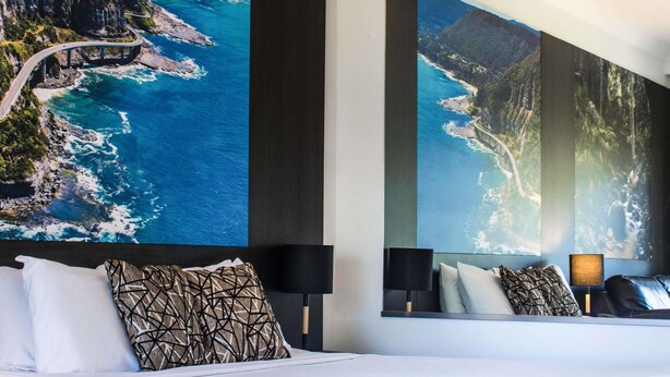 Habitación de hotel con una cama grande, una mesita de noche, una lámpara y una pared decorada con una fotografía panorámica.