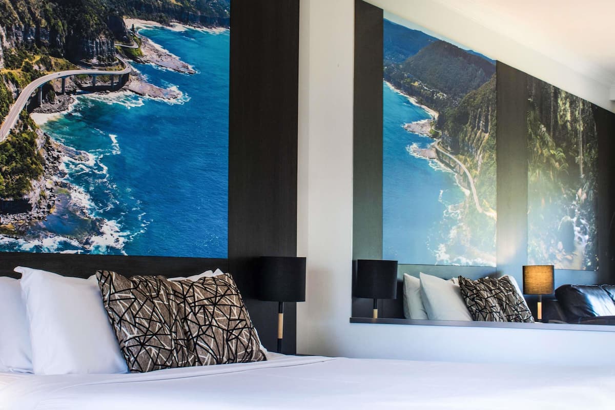 A hotel room with a large bed, a nightstand, a lamp, and a wall with a large scenic photograph.