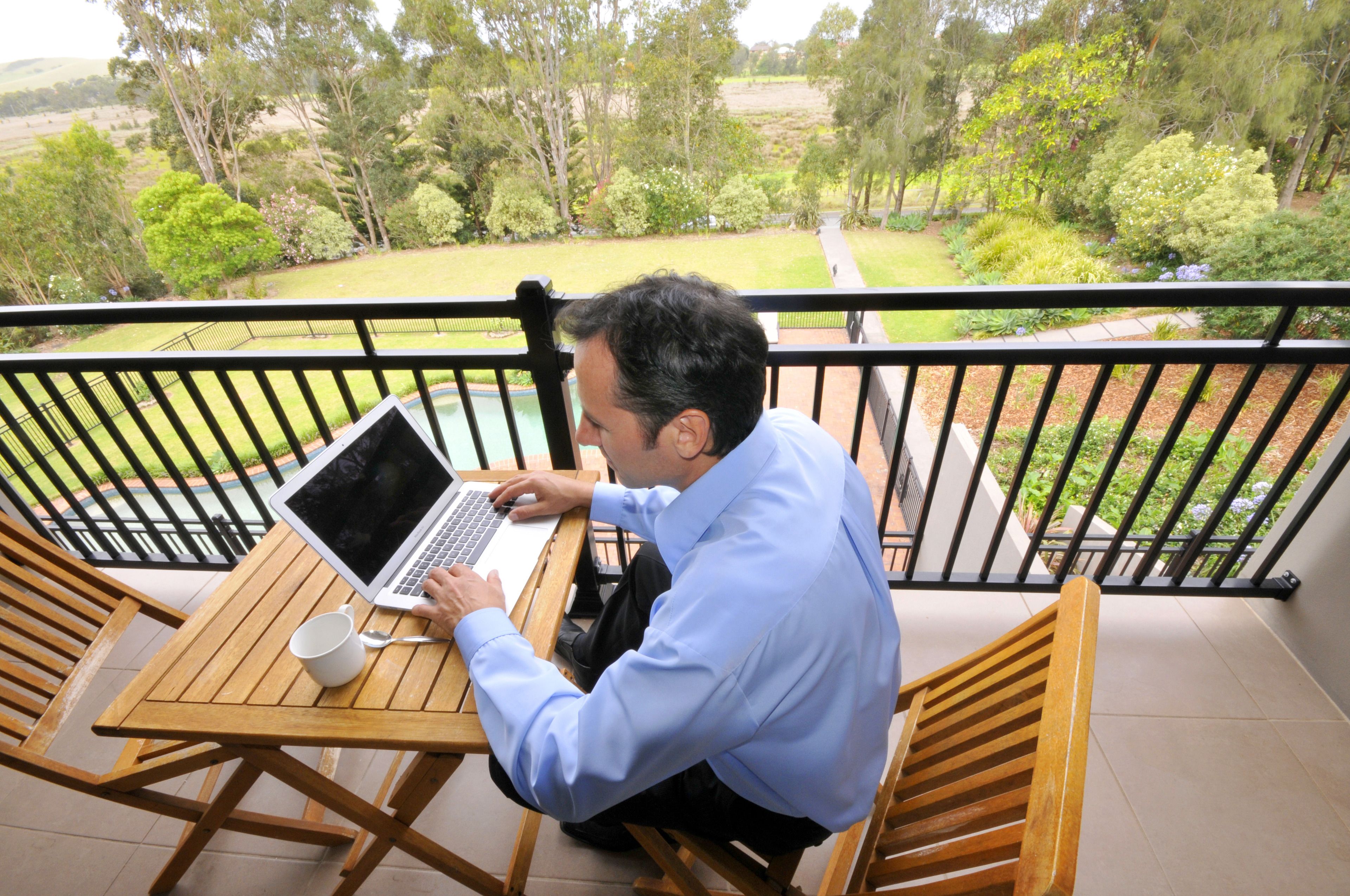 Un homme est assis à une table en bois, un ordinateur portable posé devant lui, sur un balcon donnant sur un jardin.