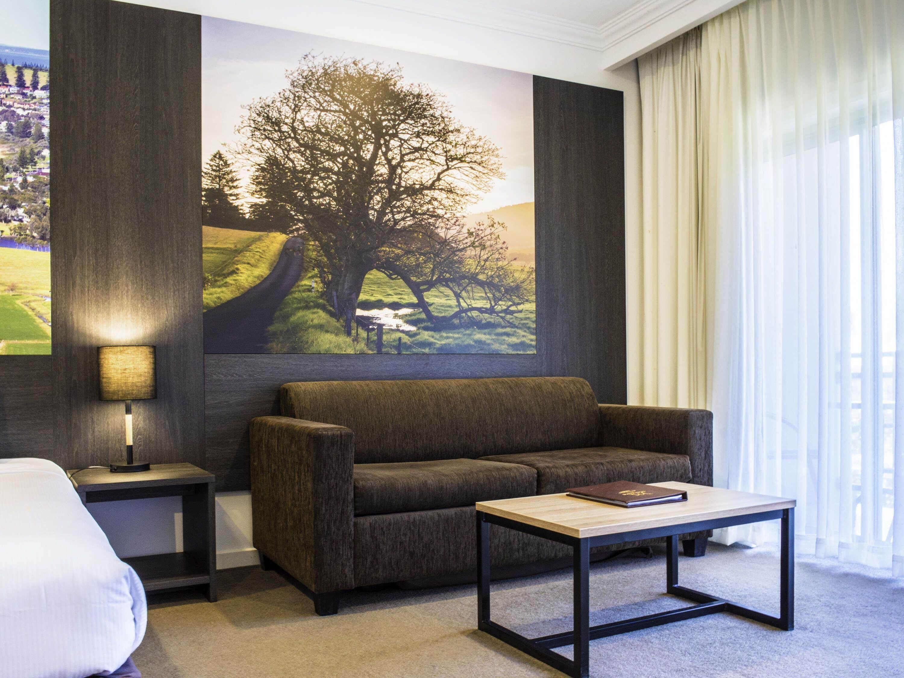 A hotel room with a sofa, a small table, a lamp, and a large painting of a tree on the wall.