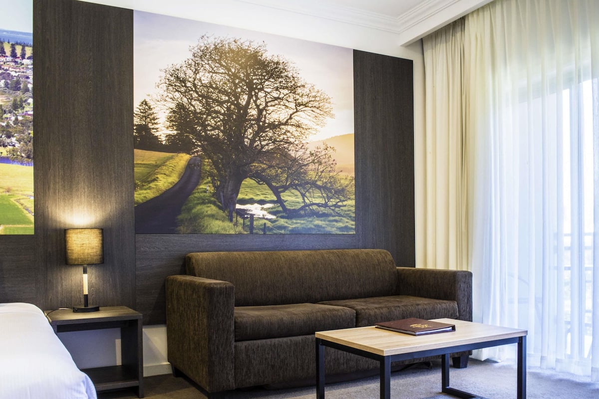 A hotel room with a sofa, a small table, a lamp, and a large painting of a tree on the wall.