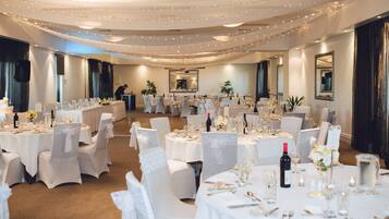 Een feestzaal met meerdere ronde tafels gedekt voor een formeel evenement, met witte tafellakens en stoelen met witte linten.