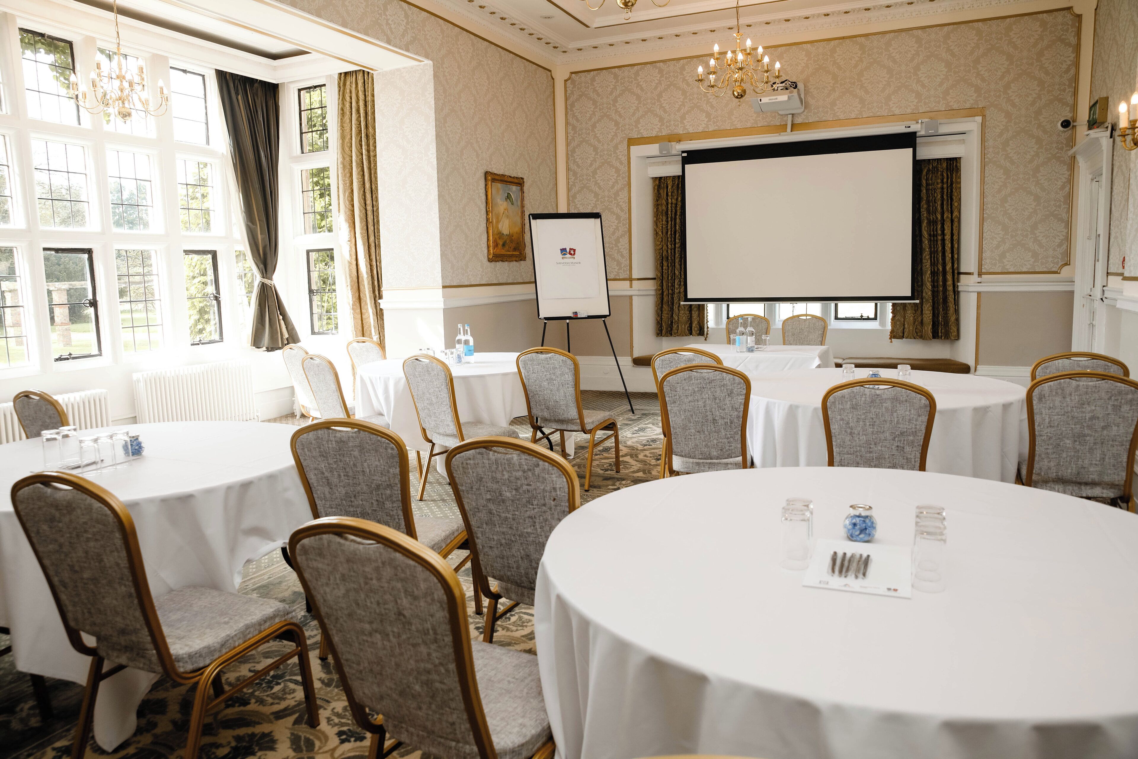 A conference room with round tables, grey upholstered chairs, a projector screen, and a whiteboard.