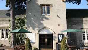 A stone building with a central entrance, two windows on the upper floor, and a small circular window above the entrance. There are outdoor tables and chairs with umbrellas, and a tree to the left. - Shendish Manor Hotel & Golf Course (Hemel Hempstead)