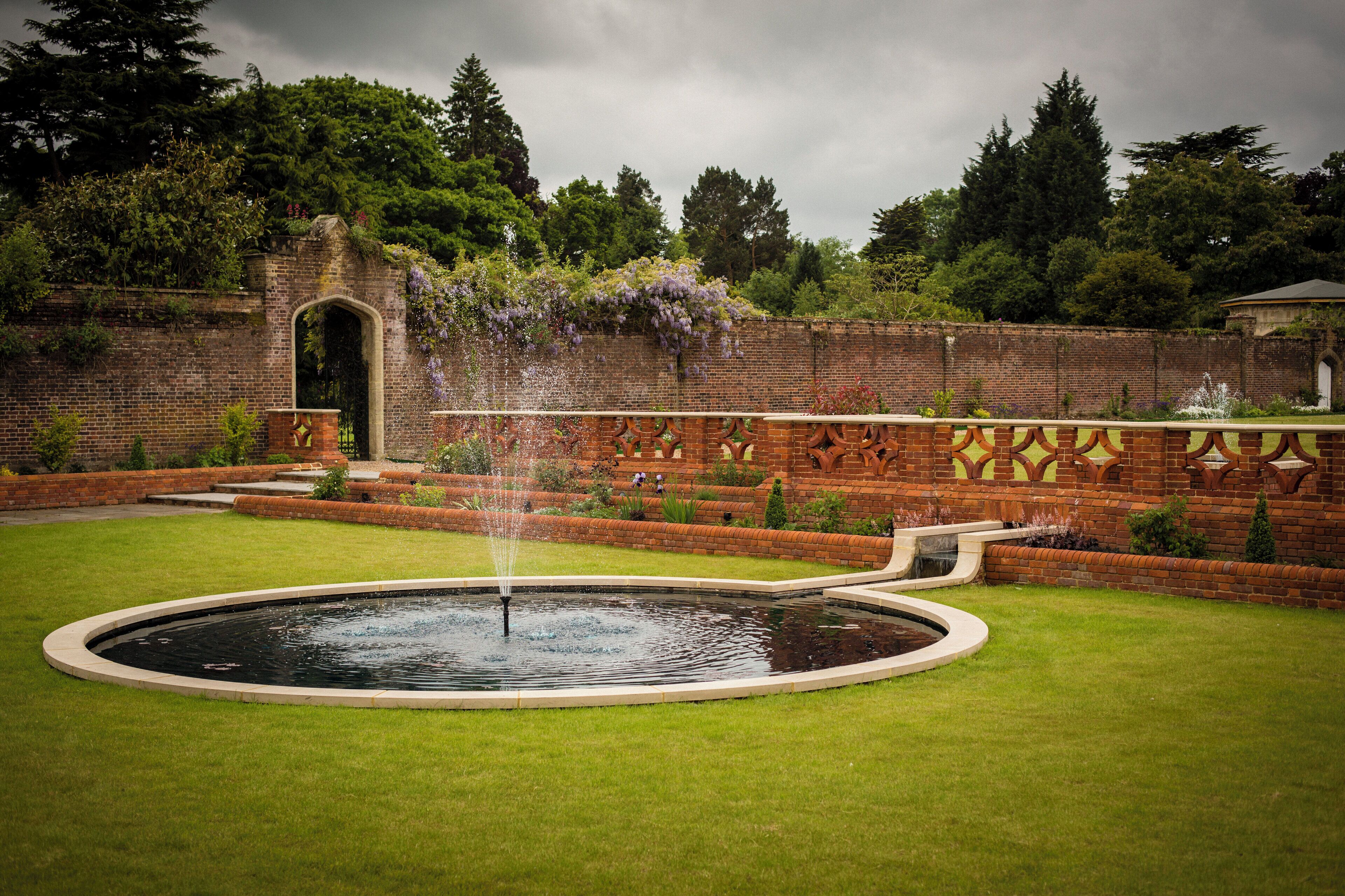 A circular fountain with water jets in a well-manicured lawn, surrounded by brick walls with decorative patterns and greenery.