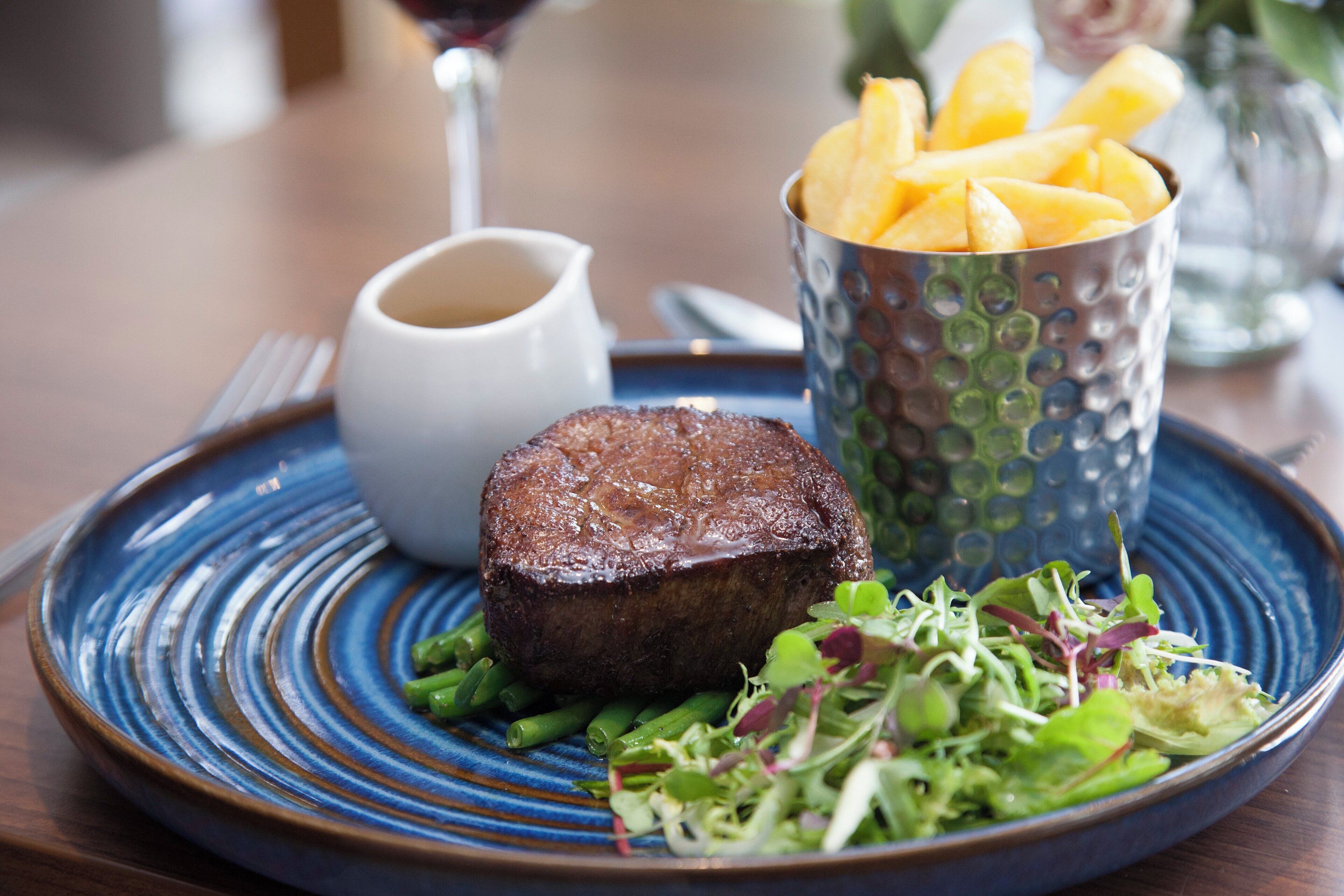 A plate with a steak, green beans, and a salad, accompanied by a cup of fries and a small pitcher of sauce.