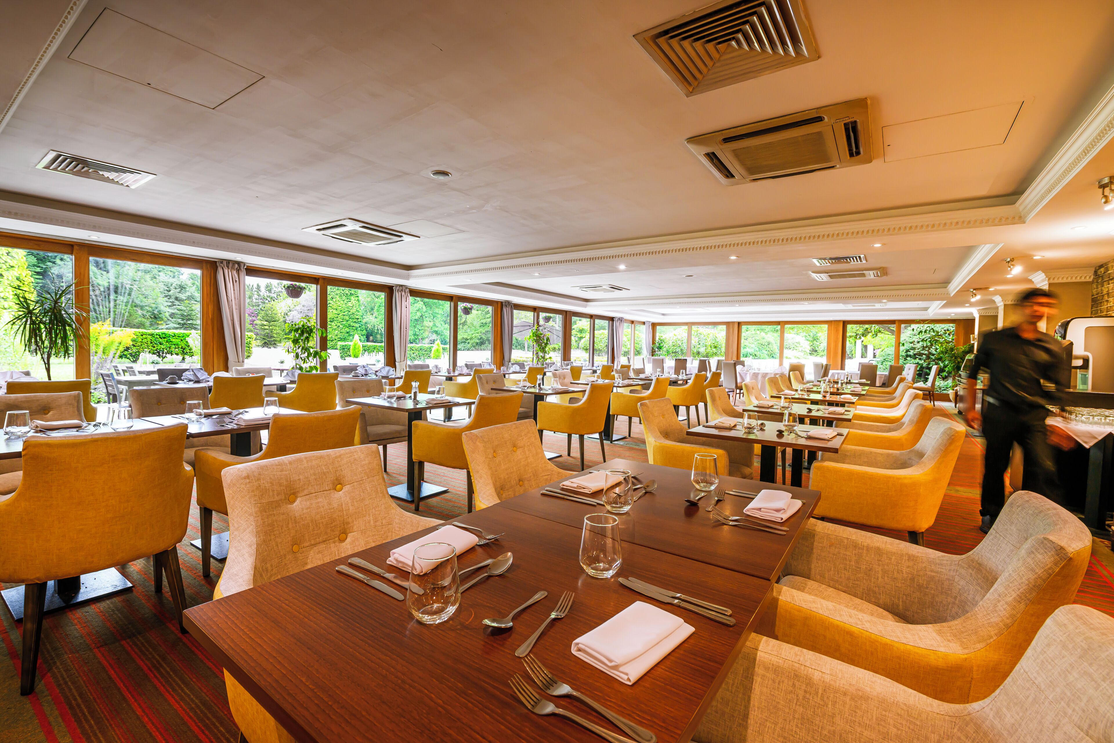 Une salle à manger d’hôtel avec de nombreuses tables et chaises, de grandes fenêtres et une vue sur la verdure environnante.