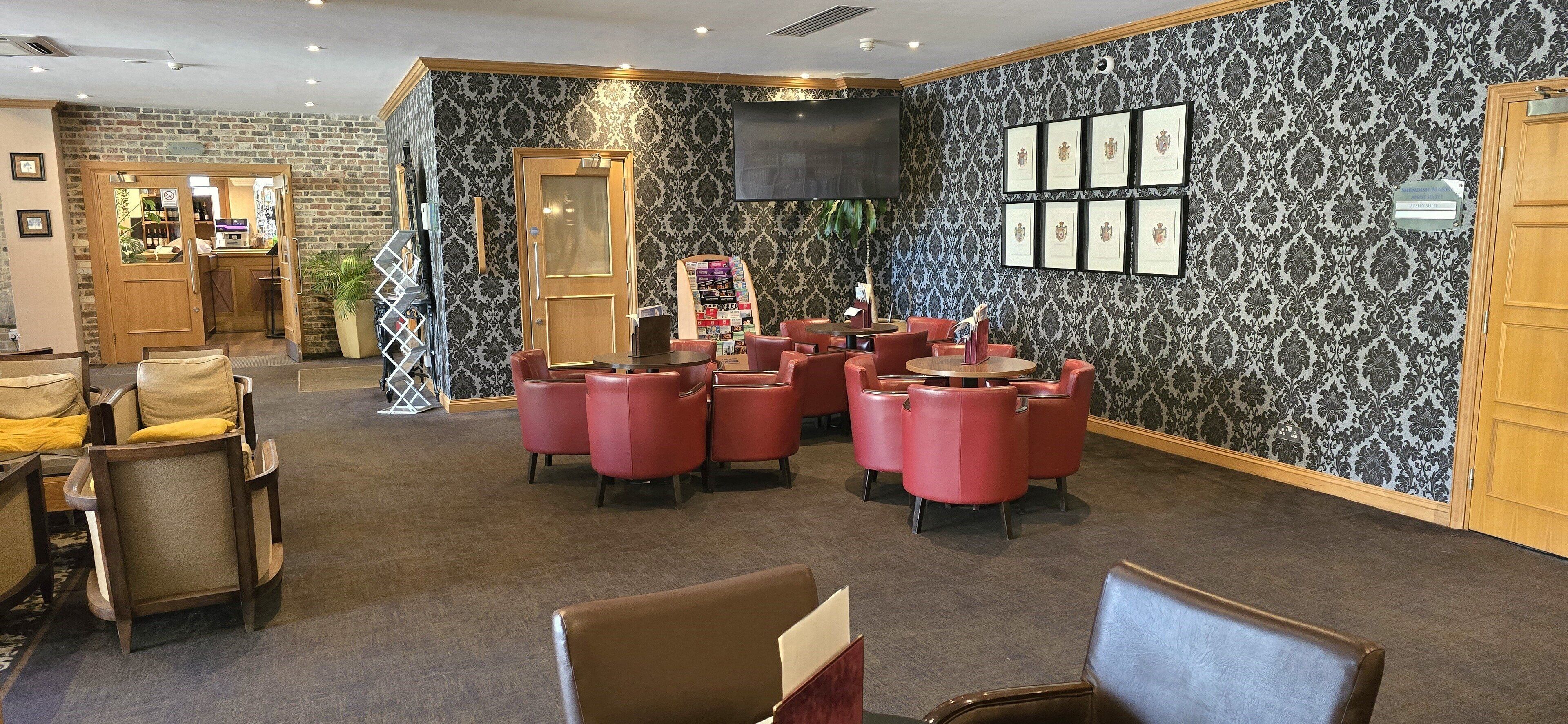 Un hall de hotel con papel tapiz estampado, sillas de cuero rojo, un mostrador de recepción y certificados enmarcados en la pared.