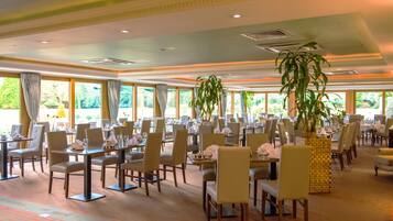 Un comedor amplio con varias mesas y sillas, ventanas grandes con cortinas y plantas en macetas.
