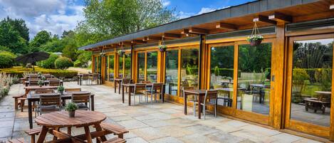 An outdoor patio area with wooden tables and chairs, large windows, and a view of the surrounding garden.