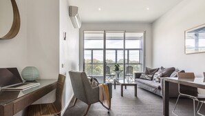Un salon moderne avec un ordinateur portable posé sur un bureau, un fauteuil gris, un canapé et une grande fenêtre donnant sur l’extérieur.