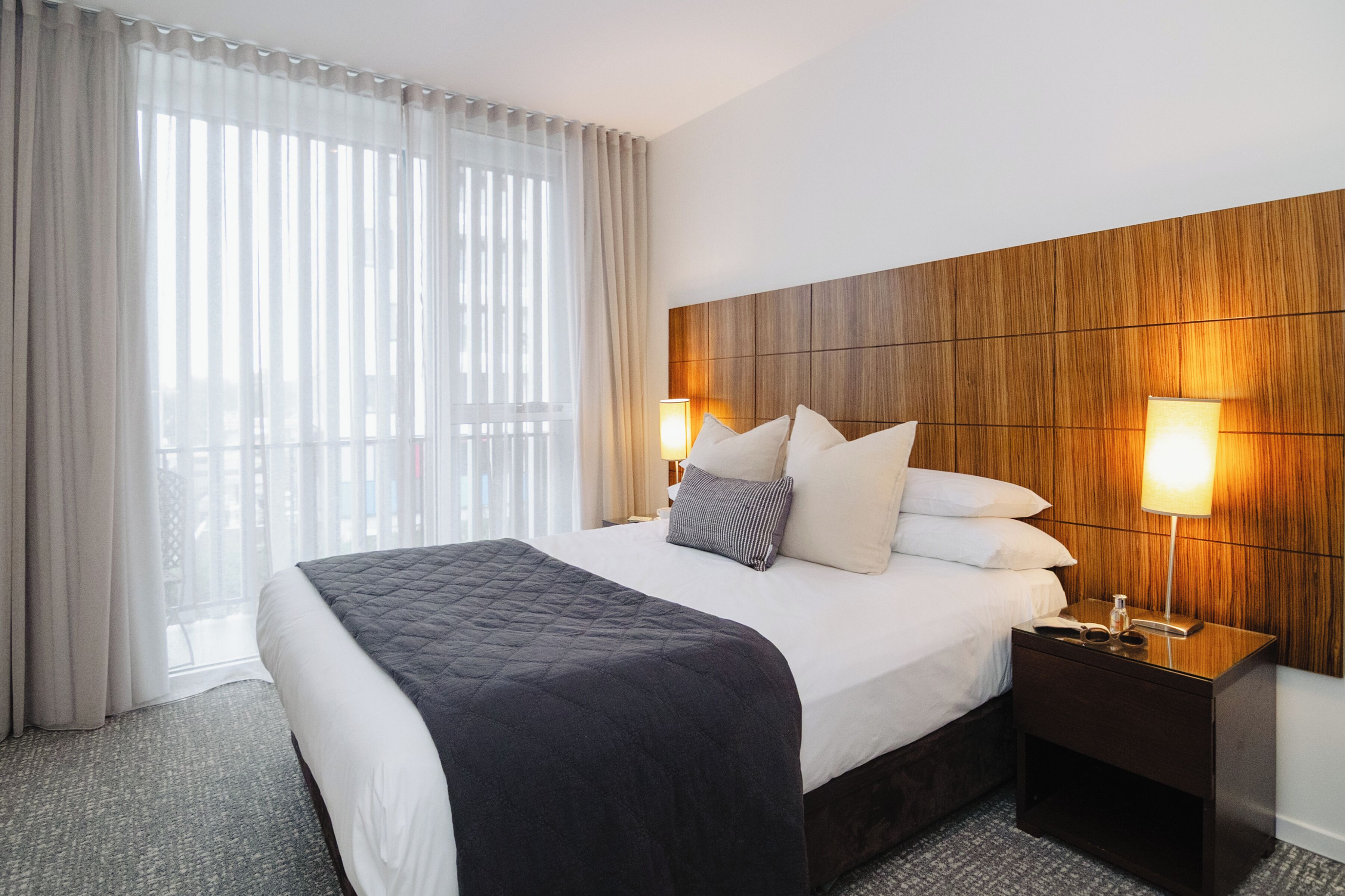 Habitación de hotel con una cama grande, dos lámparas de noche, un cabecero de madera y vistas a través de cortinas translúcidas.